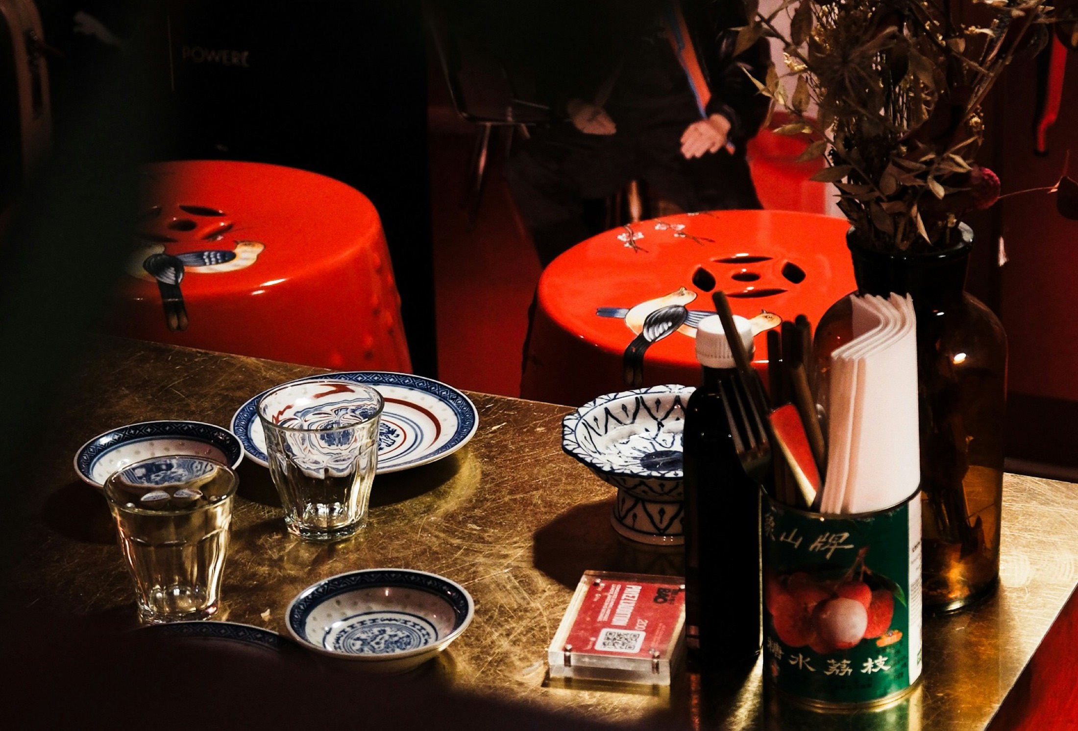 A dimly lit Chinese restaurant table set with blue-and-white porcelain bowls and glasses, red stools behind